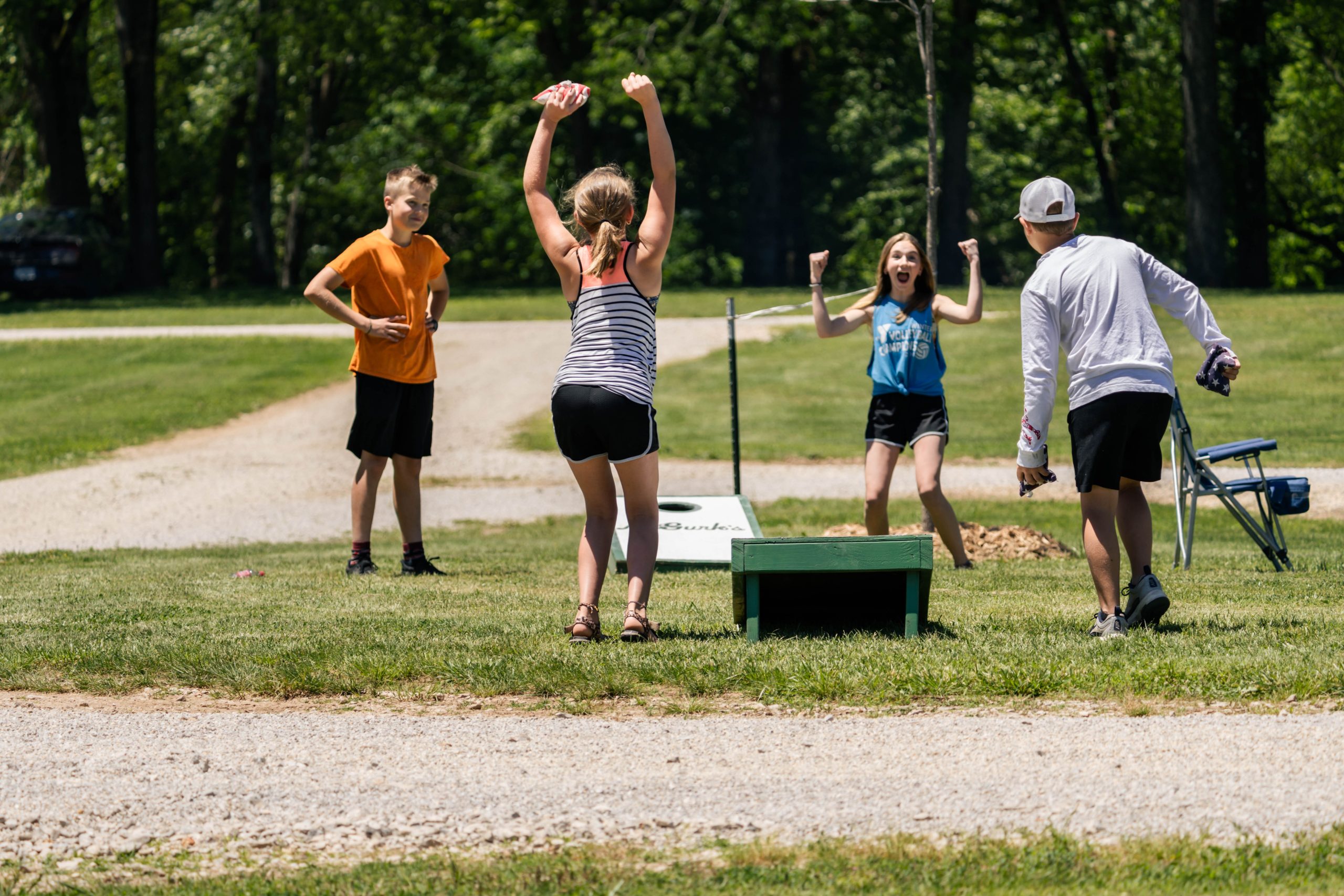 Four children are outdoors on a grassy area playing a game of cornhole. One girl is cheerfully raising her arms in victory while the other three children watch, some with their own reactions of excitement and surprise. Trees and a dirt path are visible in the background.