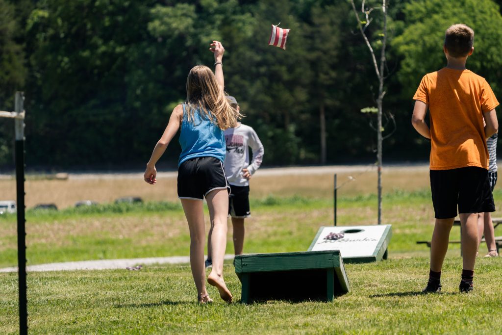 A girl tosses a bean bag towards a cornhole board in a grassy outdoor setting. She is barefoot and dressed in a blue tank top and black shorts. Several other people stand nearby, and trees provide a green backdrop.