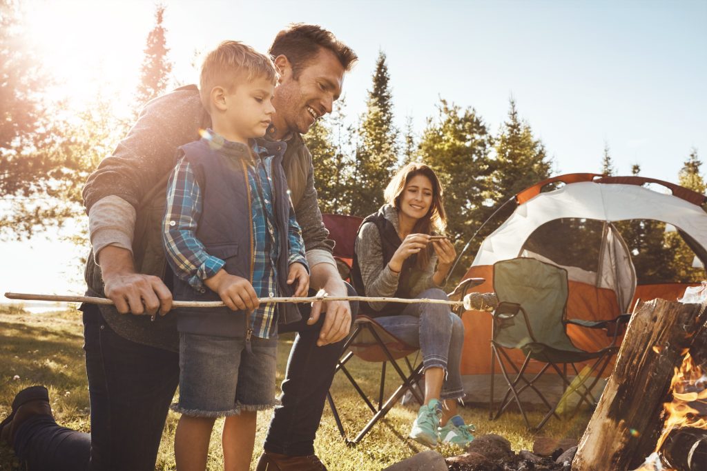 A family enjoying a camping trip outdoors. A father and his young son are roasting marshmallows over a campfire while a mother smiles, sitting in a chair near a tent. The sun is shining, and there are trees in the background.