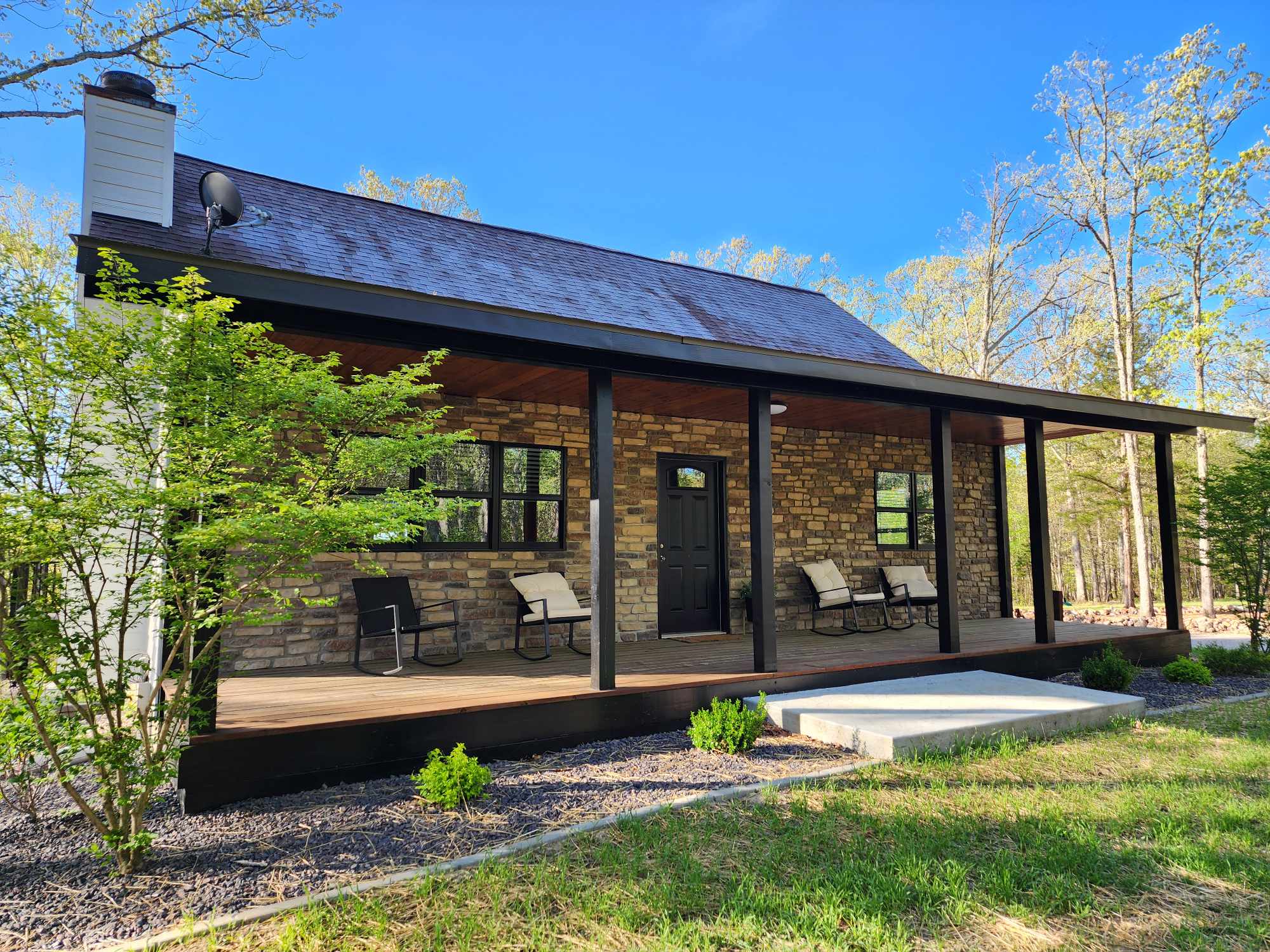 A charming stone cabin with a covered wooden porch furnished with several chairs. The porch is supported by black pillars, and the cabin is surrounded by greenery and young trees. The sky is clear and blue, suggesting a sunny day.