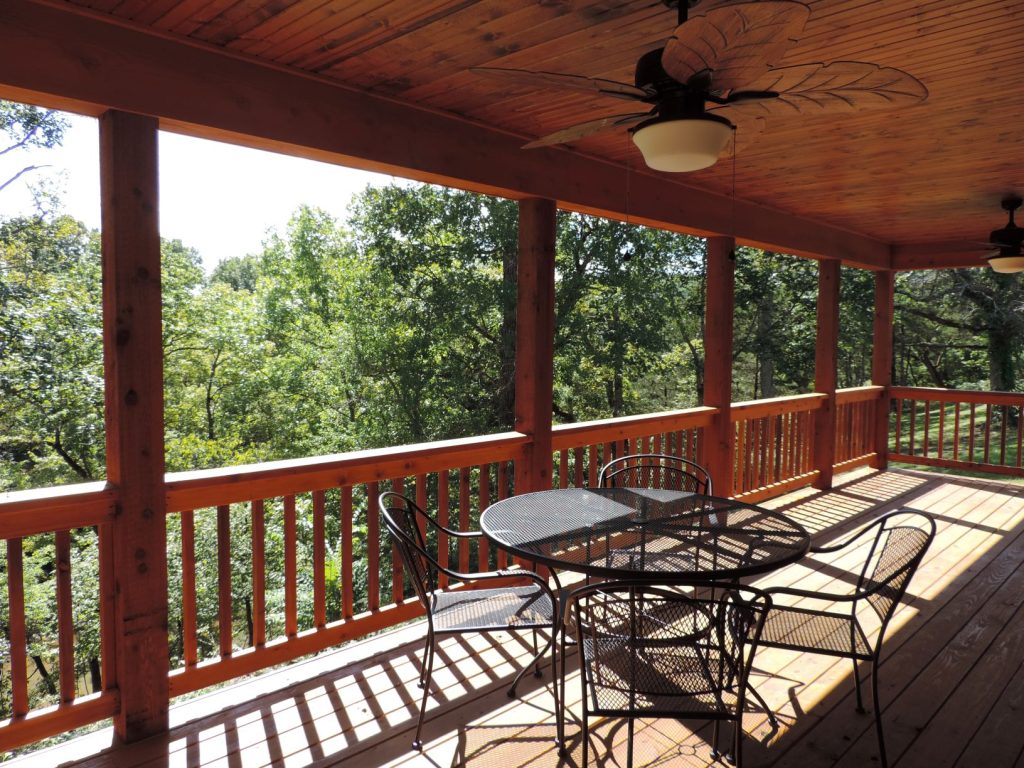 A spacious wooden deck with a ceiling fan and a round metal patio table surrounded by four chairs. The deck is shaded by a wooden roof and overlooks a lush, green forest. Sunlight streams through the rails, casting shadows on the deck floor.