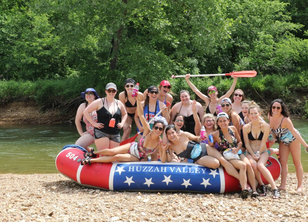 A group of people are gathered around and on top of a red, white, and blue raft labeled "HUZZAH VALLEY" on a rocky riverside. They are wearing swimsuits and sunglasses, smiling and holding paddles. Trees and a river are visible in the background.