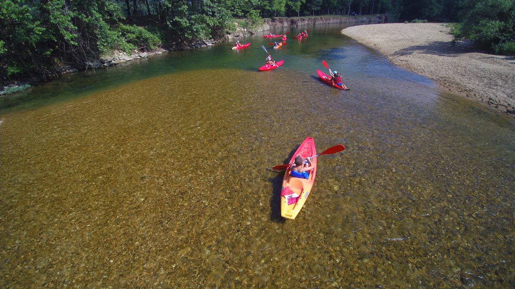 Aerial view of a group of kayakers paddling down a clear, shallow river surrounded by lush green trees. The water is transparent, revealing a rocky riverbed, and there is a sandy shore on the right side of the image.