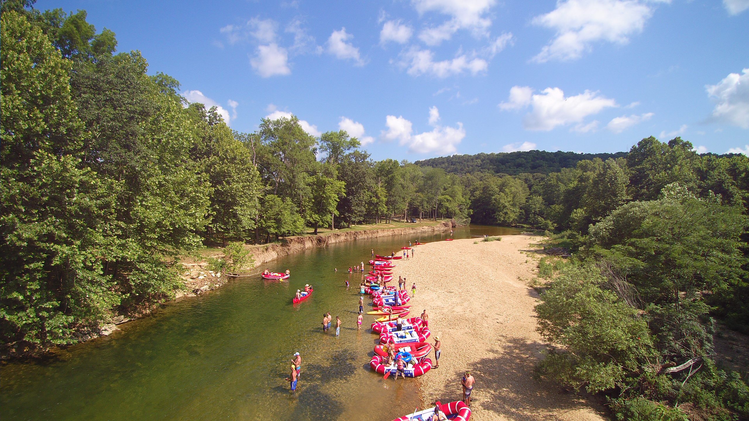 Aerial view of a clear, shallow river with people lounging on red and blue floats and swimming near a sandy beach, surrounded by lush, green trees and hills under a blue sky with scattered clouds.