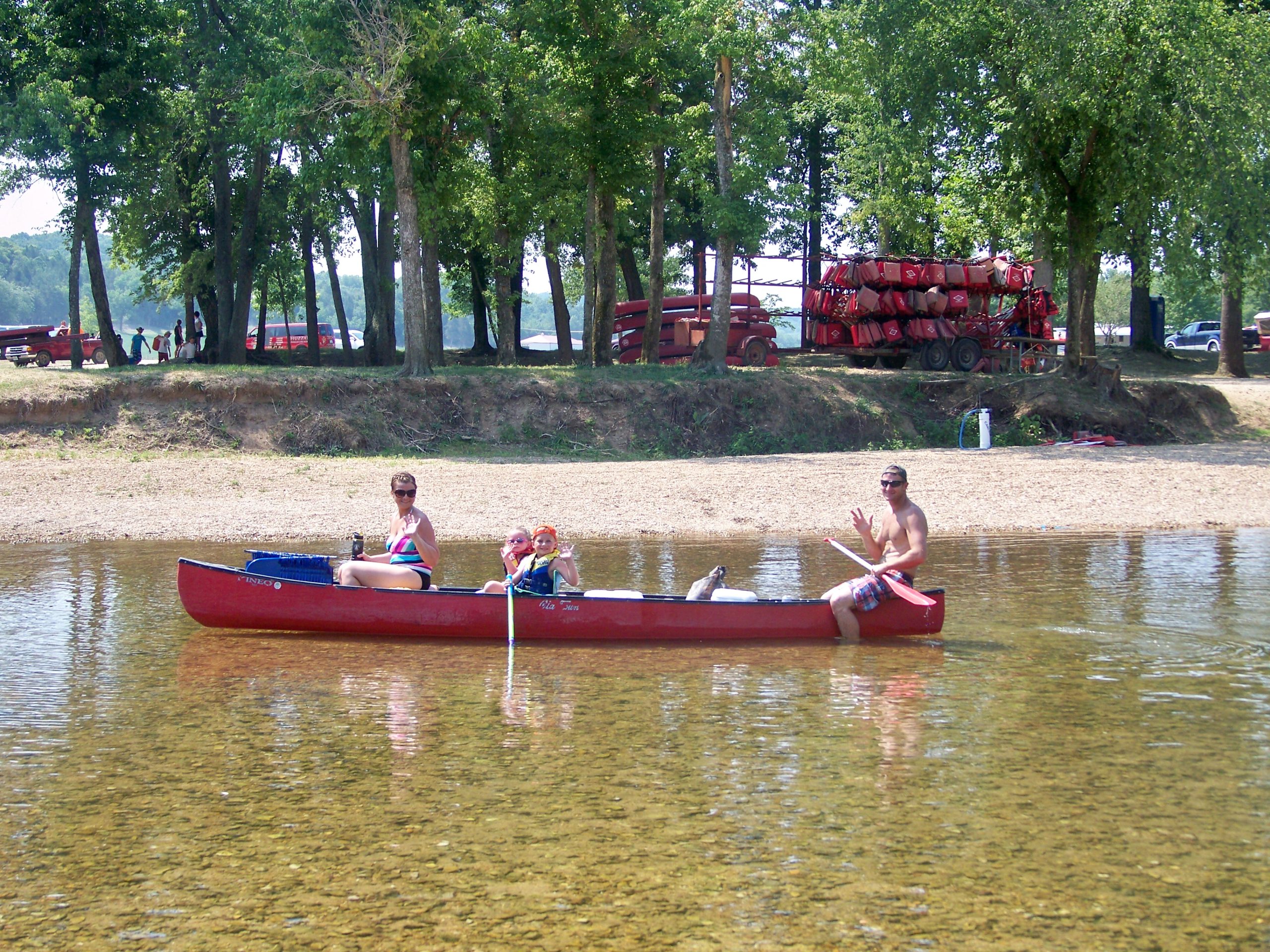 Three people enjoying a canoe ride on a calm river near a tree-lined shore. A woman, a child, and a man, who is paddling, are in the red canoe. The shore has several trees and a stack of red canoes. The water is shallow and clear. All appear to be happy and relaxed.