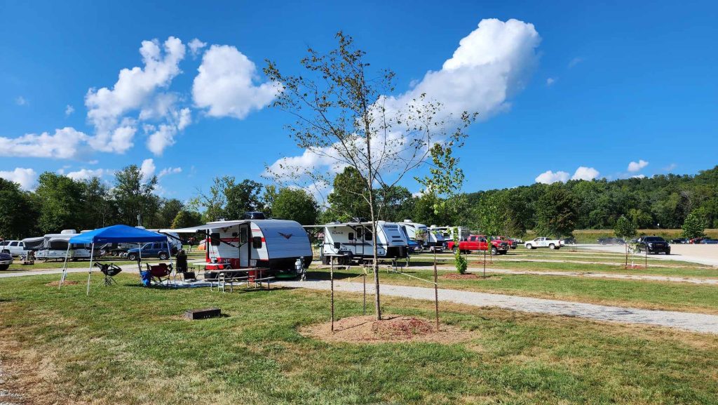 A campsite with several parked trailers under a clear blue sky. A tree stands in the foreground, surrounded by grass and dirt paths. In the background, there are trees and multiple vehicles. A blue canopy is set up near one trailer.