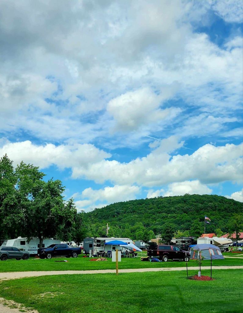 A scenic campground with several RVs and cars parked on a grassy area. Tents and picnic tables are set up under a partly cloudy sky. A forested hill is visible in the background, and an umbrella is open in the foreground.