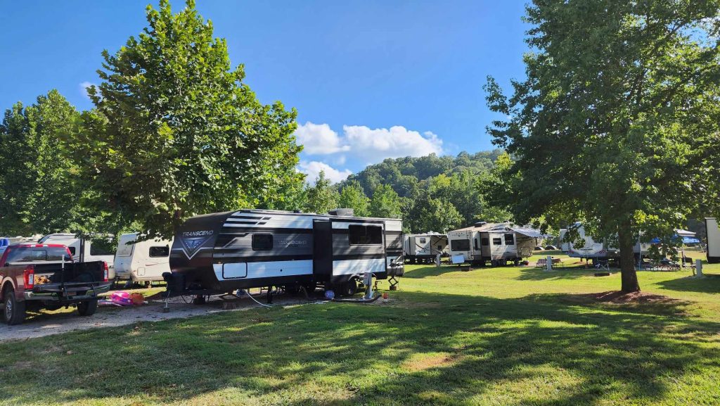 A campsite with multiple RVs parked on a grassy area surrounded by green trees. A red truck is parked beside a black and white trailer. The sky is clear with some fluffy clouds, and a hillside with trees is visible in the background.