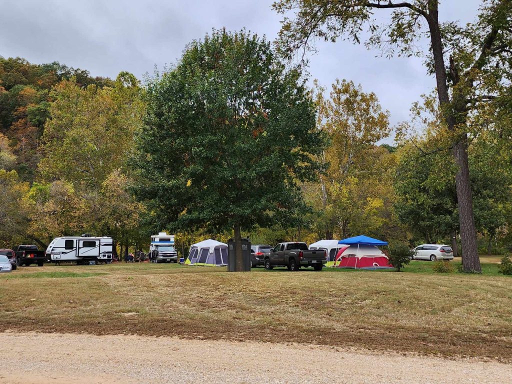 A campsite with several tents and parked vehicles among trees with autumn foliage. A grassy area is in the foreground, and the sky is overcast. A recreational vehicle and a few people are visible in the background.