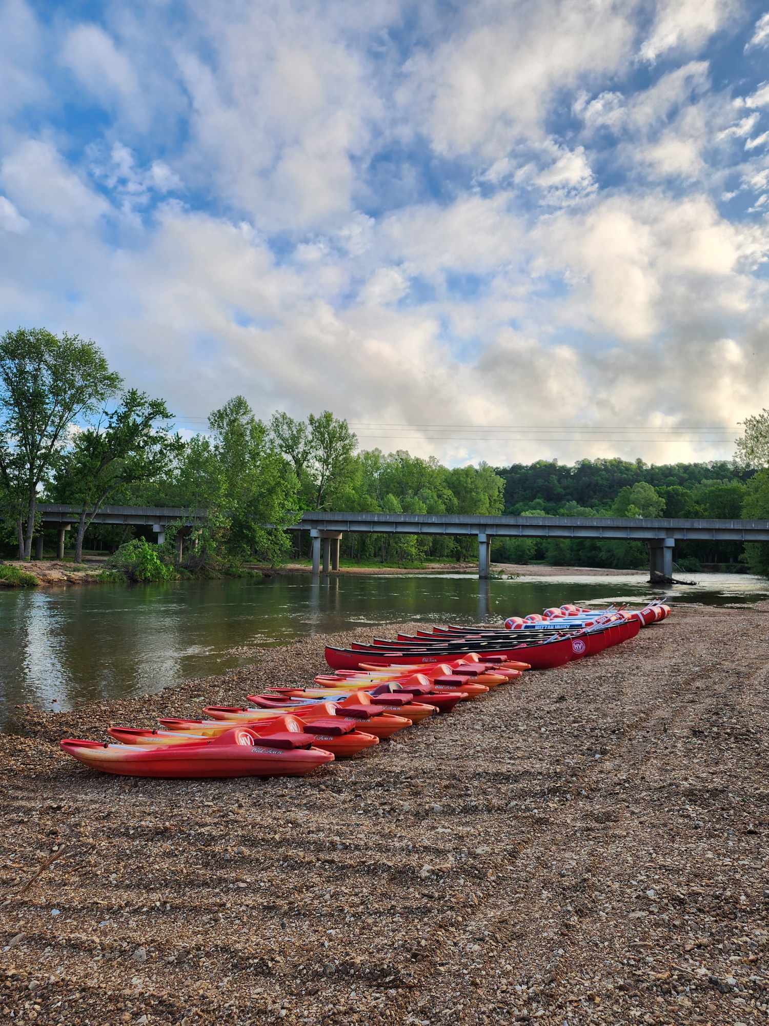 A group of red and orange canoes lined up on a pebbled shore by a calm river. The background features lush green trees and a bridge under a partly cloudy sky.