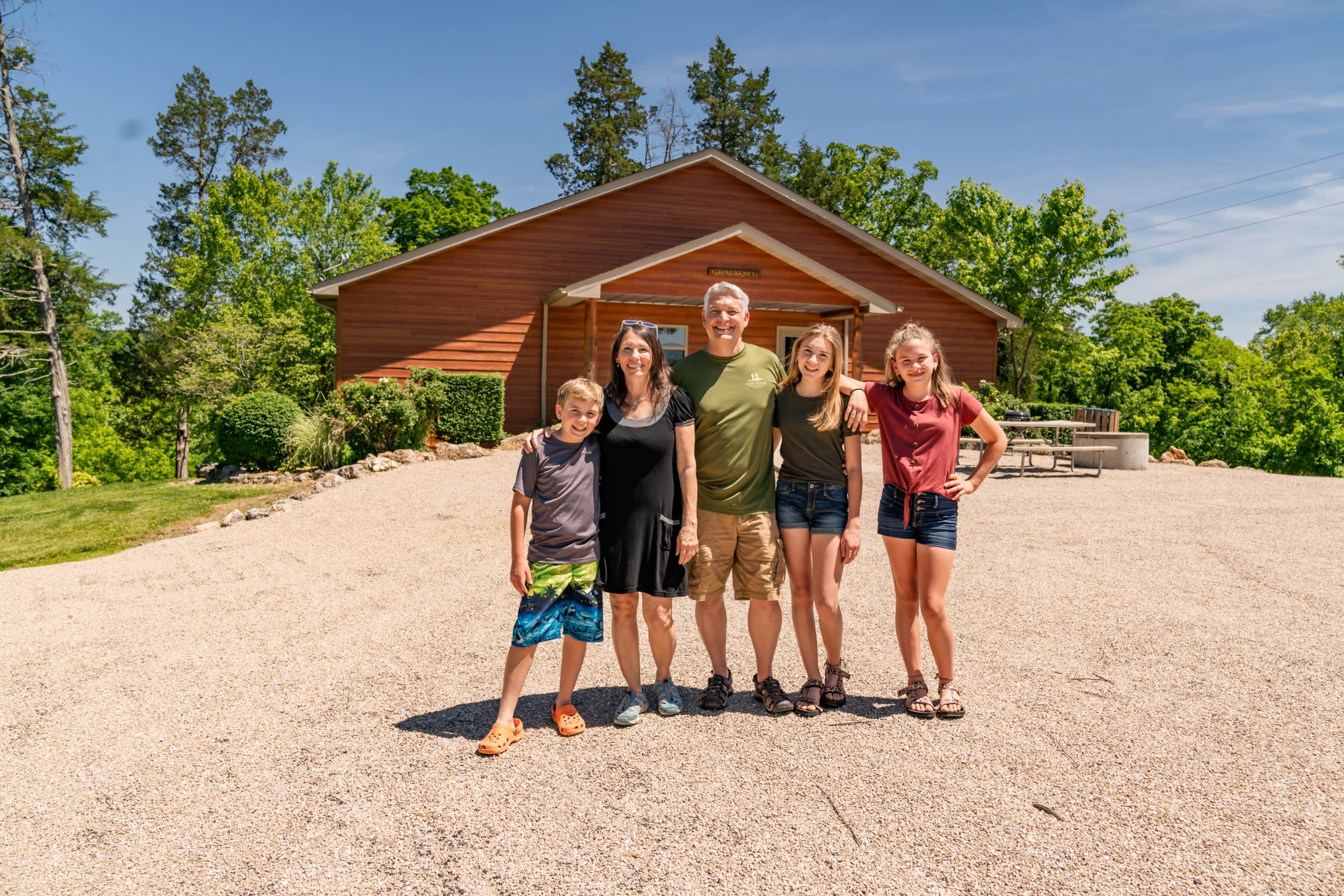 Family of five smiling in front of a wooden house with lush greenery and clear blue sky. They stand on a gravel path, dressed casually for warm weather, arms around each other.