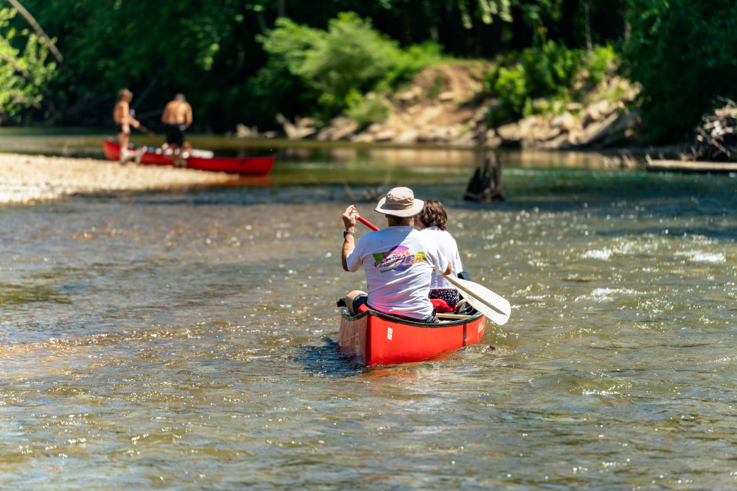 Two people paddle a red canoe on a shallow river, seen from behind. One wears a hat and holds a phone. In the background, two people in another red canoe approach the riverbank. Trees line the river, reflecting a sunny day.