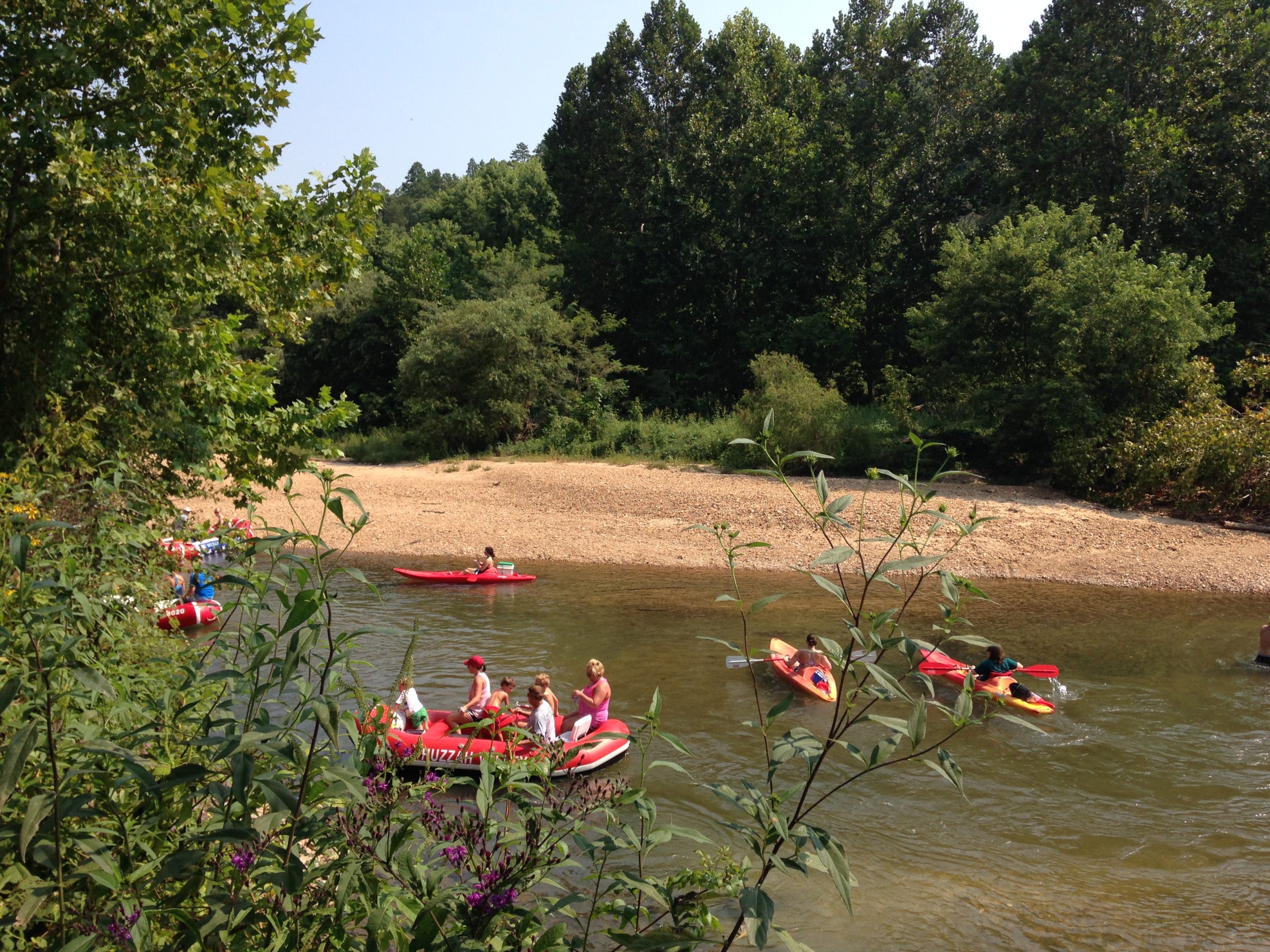 People are kayaking and rafting on a calm river near a sandy bank surrounded by lush greenery on a sunny day.