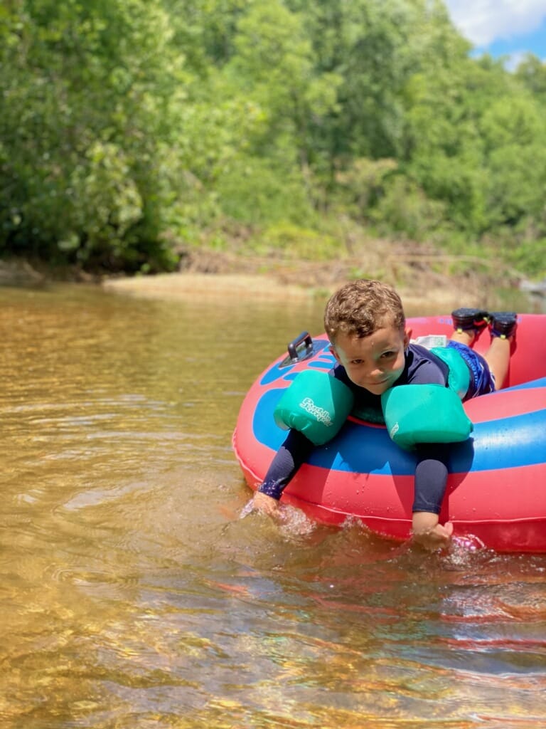 A young boy wearing a blue life vest relaxes on an inflatable red and blue raft in a shallow, clear stream. The background features lush green foliage on a sunny day.