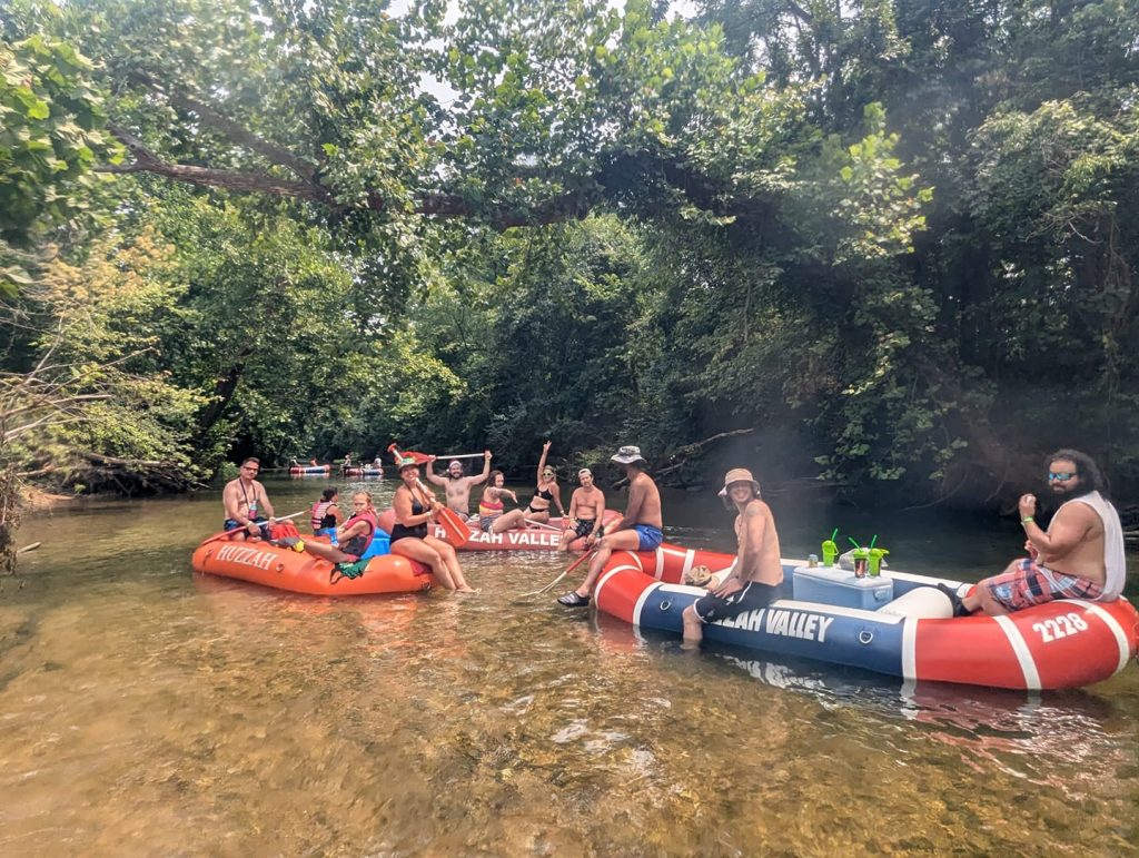 A group of people are floating down a river in colorful inflatable rafts. They are surrounded by lush green trees. Some are waving and holding drinks, enjoying a sunny day outdoors. The water is shallow and clear.