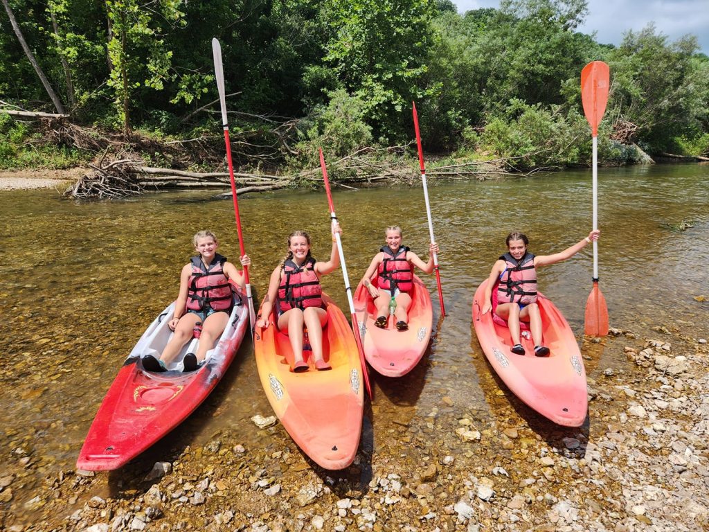 Four people in life vests sit in red kayaks on a clear, shallow river with paddles raised. They are near a rocky shore, surrounded by green trees and vegetation. The sky is partly cloudy.